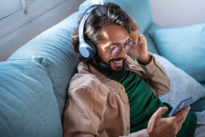 Smiling young man wearing headphones listen music playing in smartphone sitting on sofa at home
